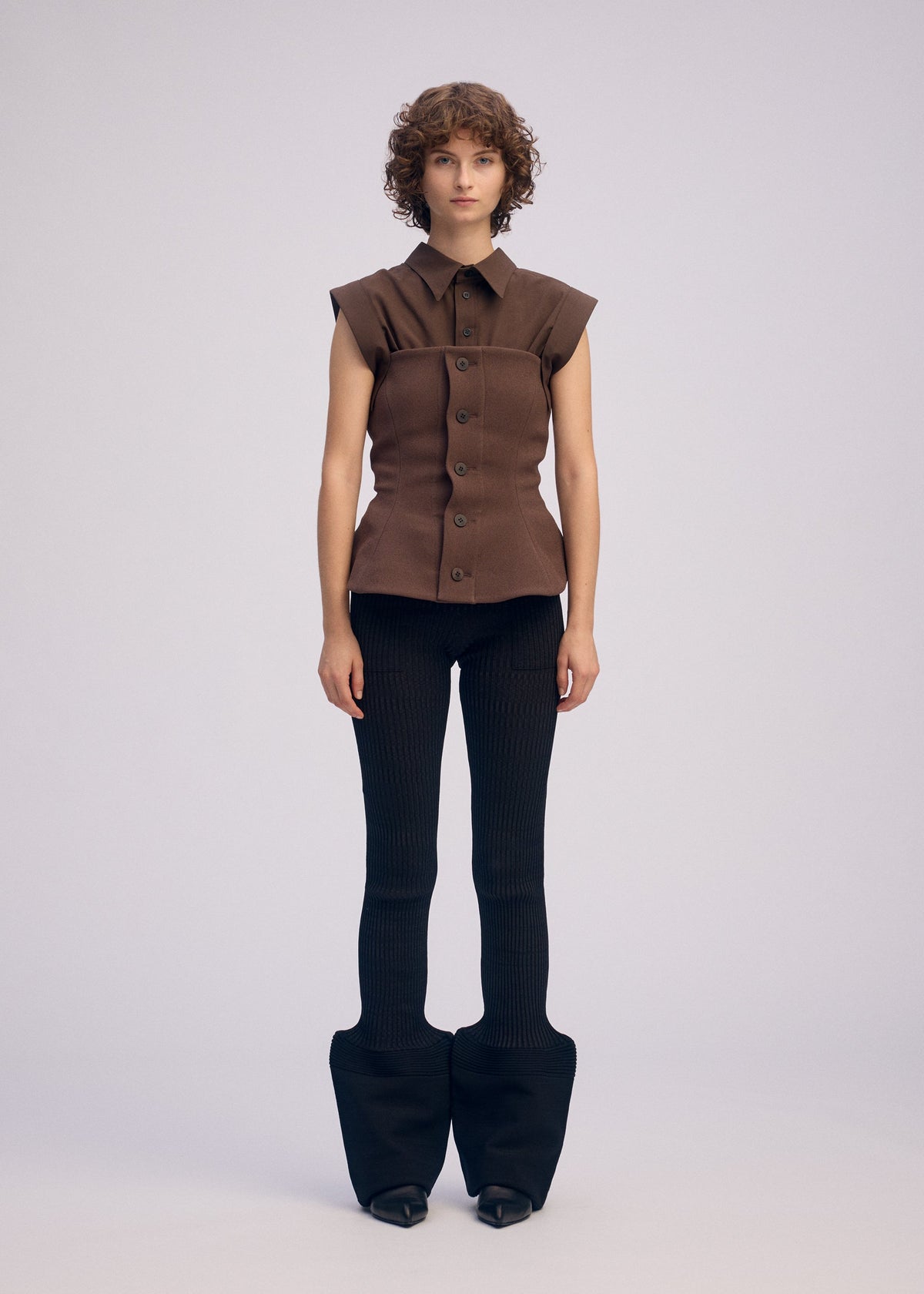 A person with short curly hair wears the ARMS sleeveless brown bustier button-up top by ISSEY MIYAKE and fitted black flared pants. The background is plain light gray. view 5 of 7