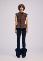A person with short curly hair wears the ARMS sleeveless brown bustier button-up top by ISSEY MIYAKE and fitted black flared pants. The background is plain light gray.