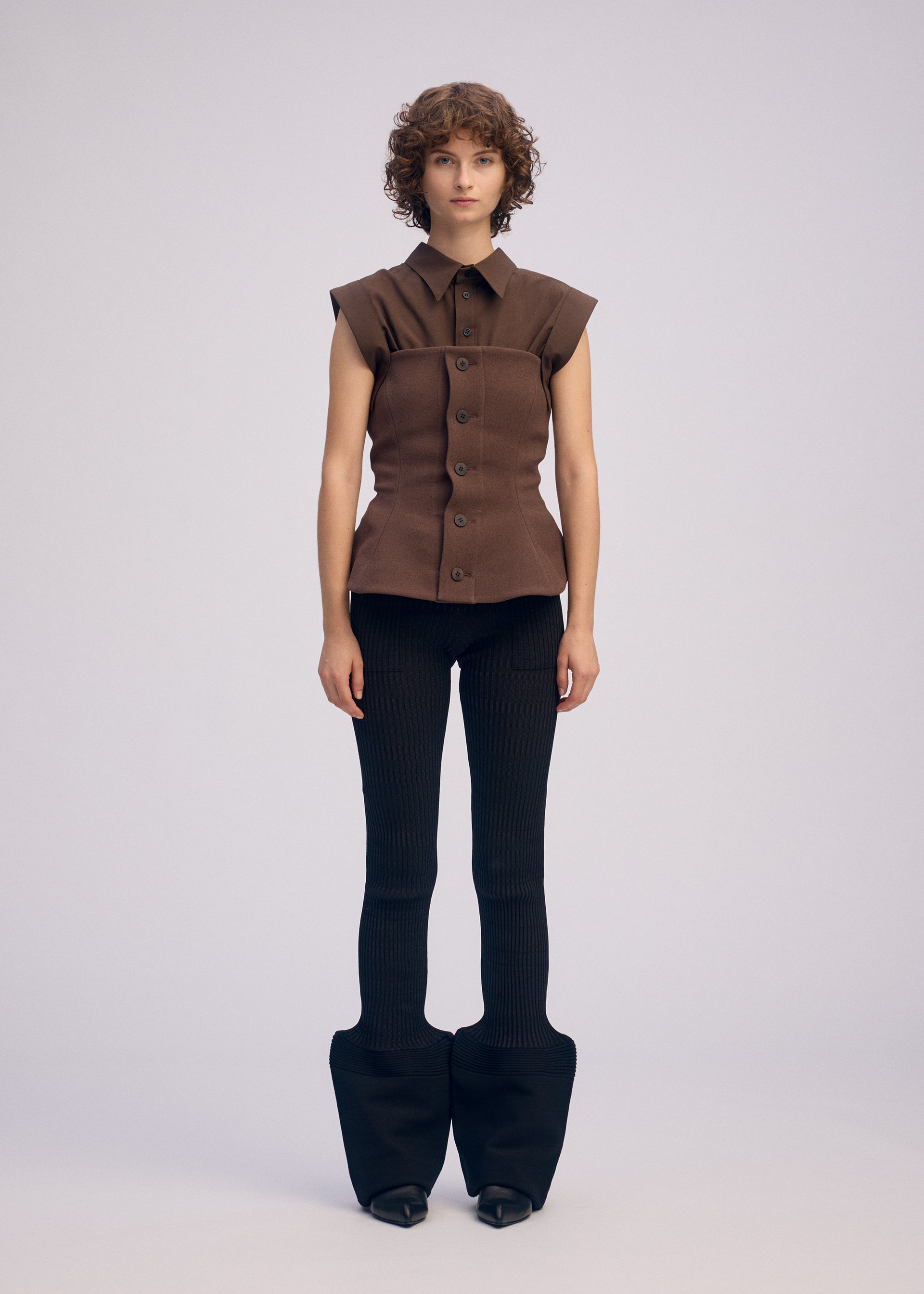 Zoomed view A person with short curly hair wears the ARMS sleeveless brown bustier button-up top by ISSEY MIYAKE and fitted black flared pants. The background is plain light gray.
