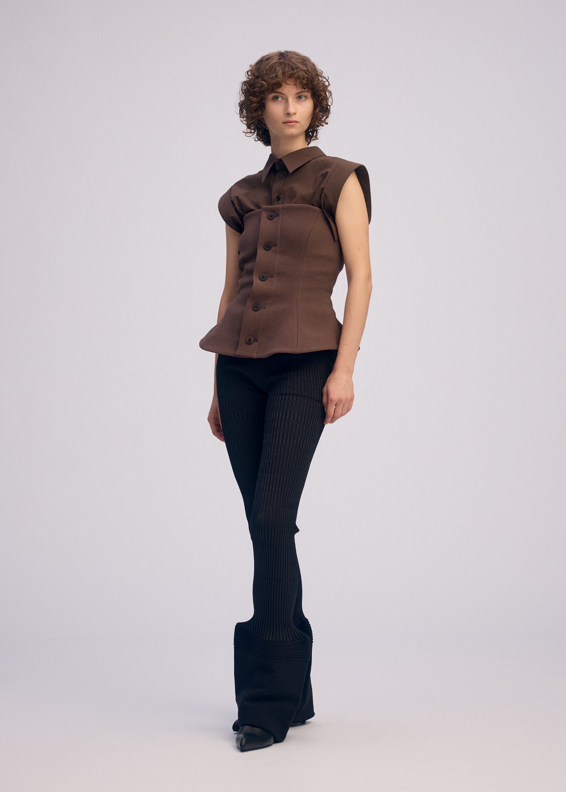 Zoomed view A person with short curly hair models the ARMS bustier-style buttoned top by ISSEY MIYAKE, featuring a curved waist and structured shoulders, paired with fitted black stretch flared pants, against a plain light background.