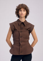 A person with short, curly brown hair wears ISSEY MIYAKE's ARMS bustier-inspired sleeveless brown top, featuring structured corset seams and a curved waist, paired with dark pants against a plain light background.