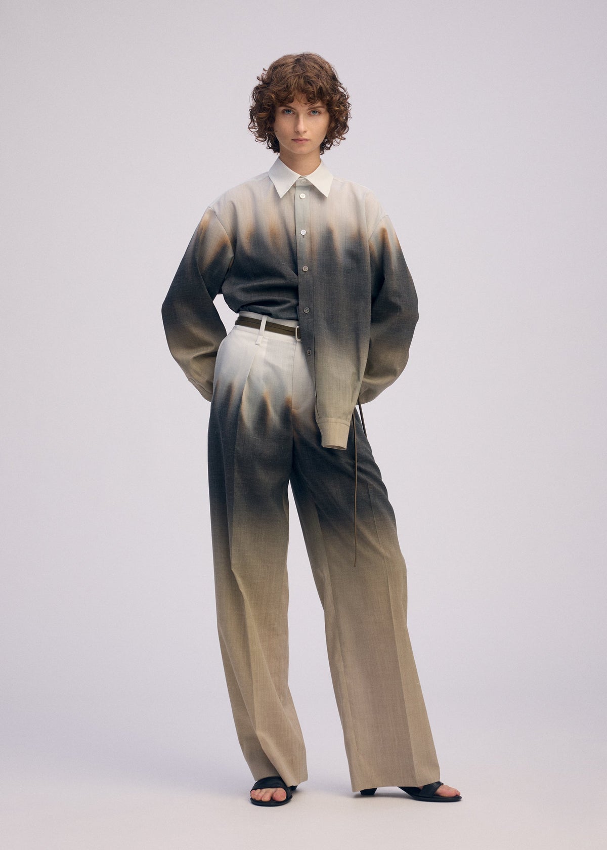 A person with curly brown hair wears ISSEY MIYAKE's PLUMAGE set—loose, gradient feather-patterned shirt and matching wide-leg trousers—styled with a white belt and black sandals against a plain light background. view 8 of 9