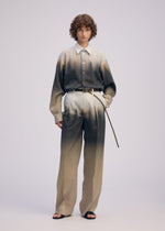 A person with short curly hair wears the ISSEY MIYAKE PLUMAGE set—an artisan Kyoto shirt and matching high-waisted trousers with a gradient feather pattern in neutral tones—paired with black sandals and a thin belt, against a plain light background.