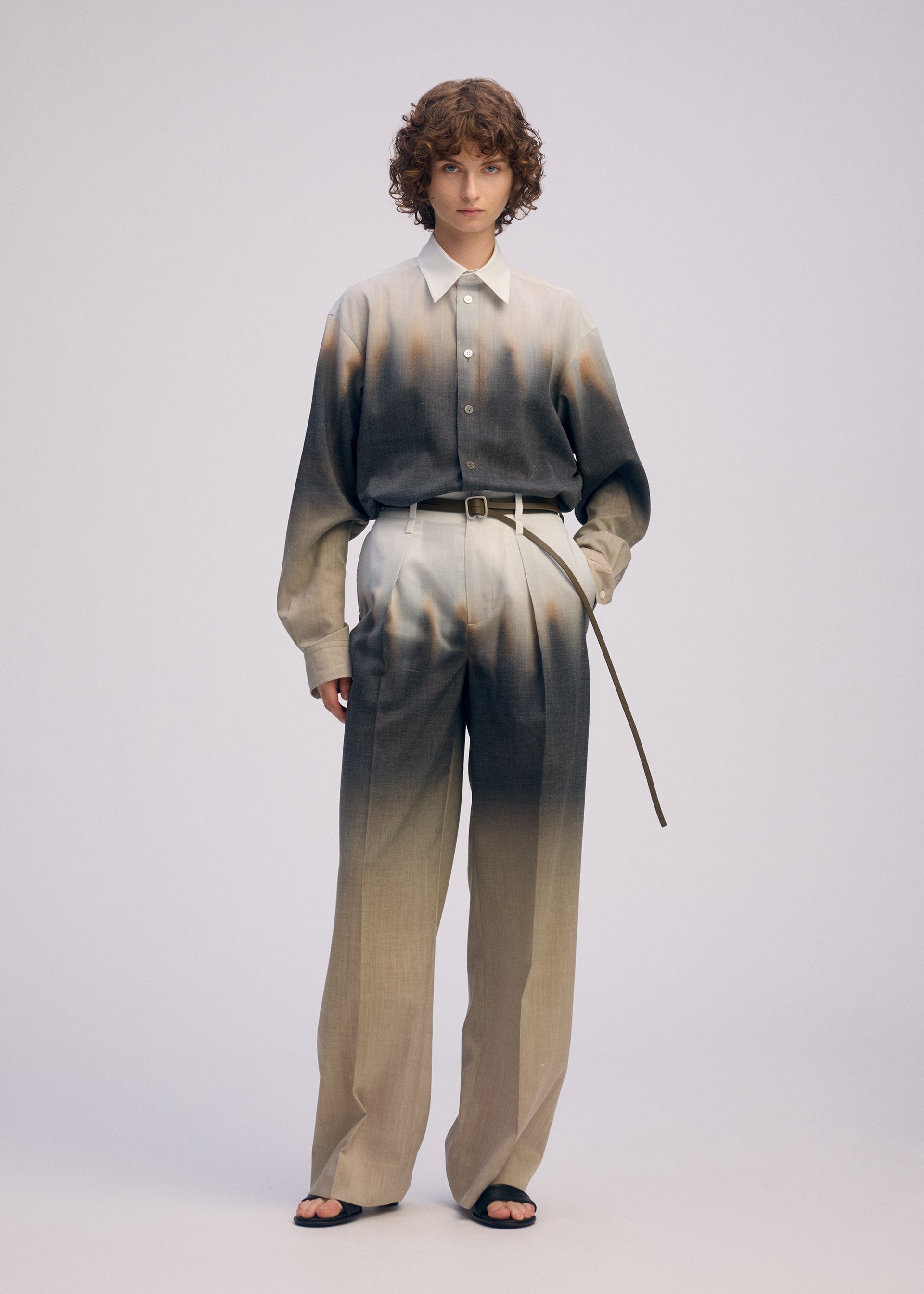 Zoomed view A person with short curly hair wears the ISSEY MIYAKE PLUMAGE set—an artisan Kyoto shirt and matching high-waisted trousers with a gradient feather pattern in neutral tones—paired with black sandals and a thin belt, against a plain light background.
