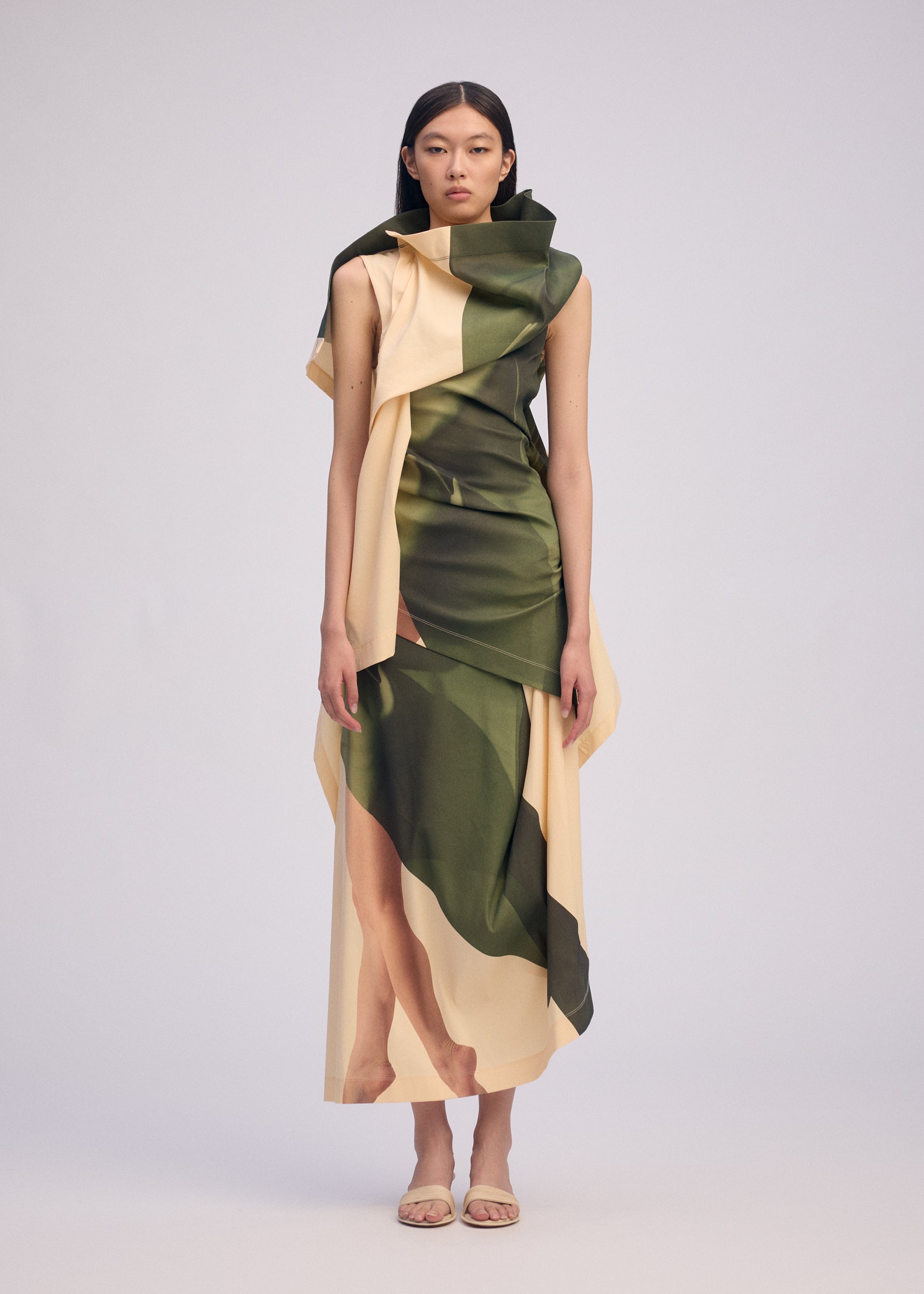 Zoomed view A woman stands in a studio wearing ISSEY MIYAKE's BODY AT PLAY, a sleeveless, asymmetrical ribbed stretch dress with bold green and beige abstract patterns. She pairs it with nude sandals against a plain light background.