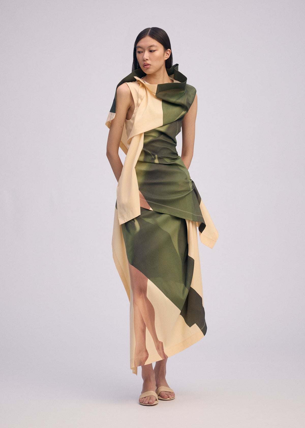 A woman poses against a plain light background wearing the BODY AT PLAY sleeveless draped dress by ISSEY MIYAKE in green and beige with an original pattern. She has long, straight dark hair and nude sandals. view 4 of 6