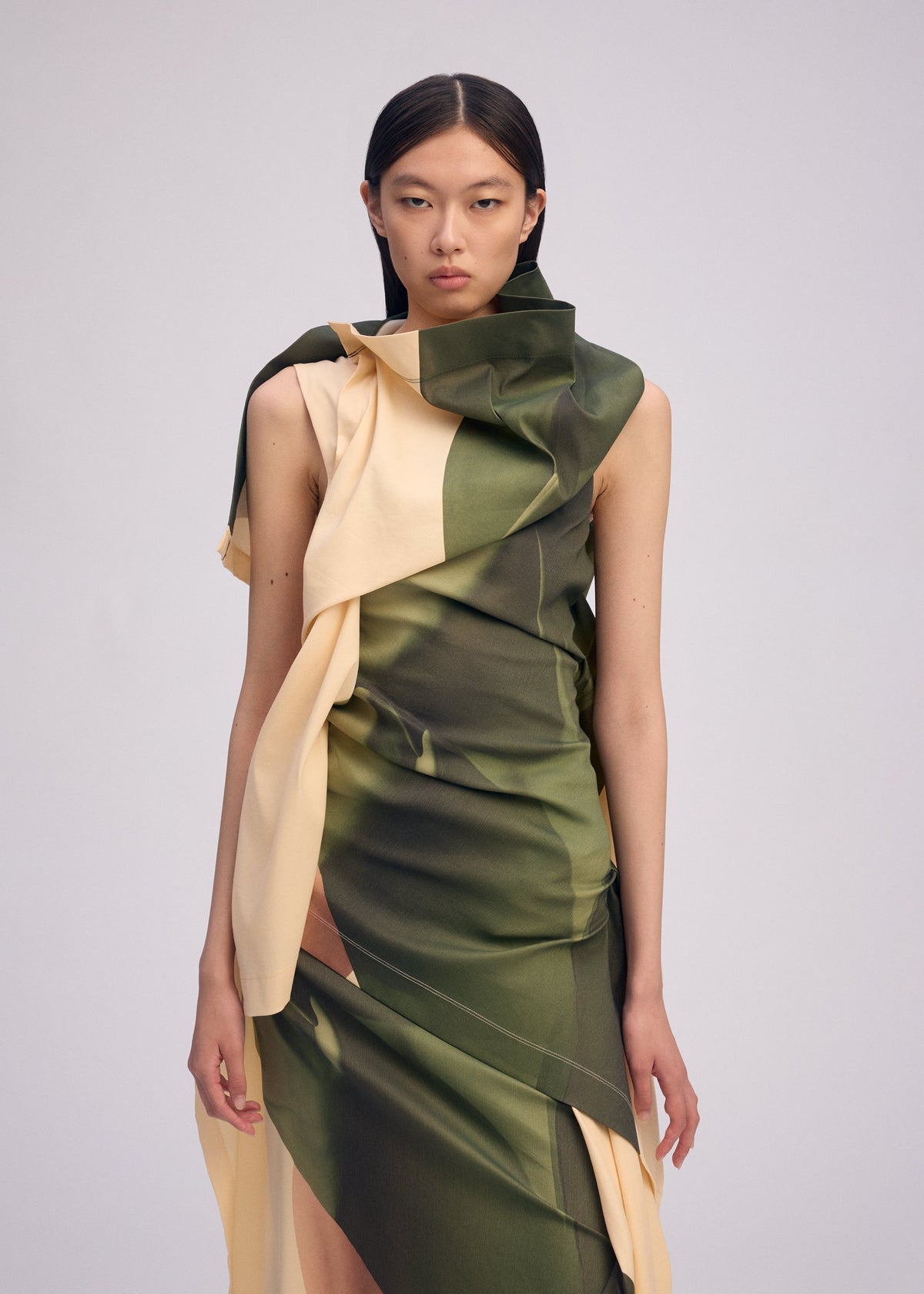A woman with straight dark hair wears BODY AT PLAY, a sleeveless draped dress with green and beige abstract patterns by ISSEY MIYAKE, standing against a plain light background. view 5 of 6