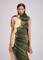 A woman with straight dark hair wears BODY AT PLAY, a sleeveless draped dress with green and beige abstract patterns by ISSEY MIYAKE, standing against a plain light background.