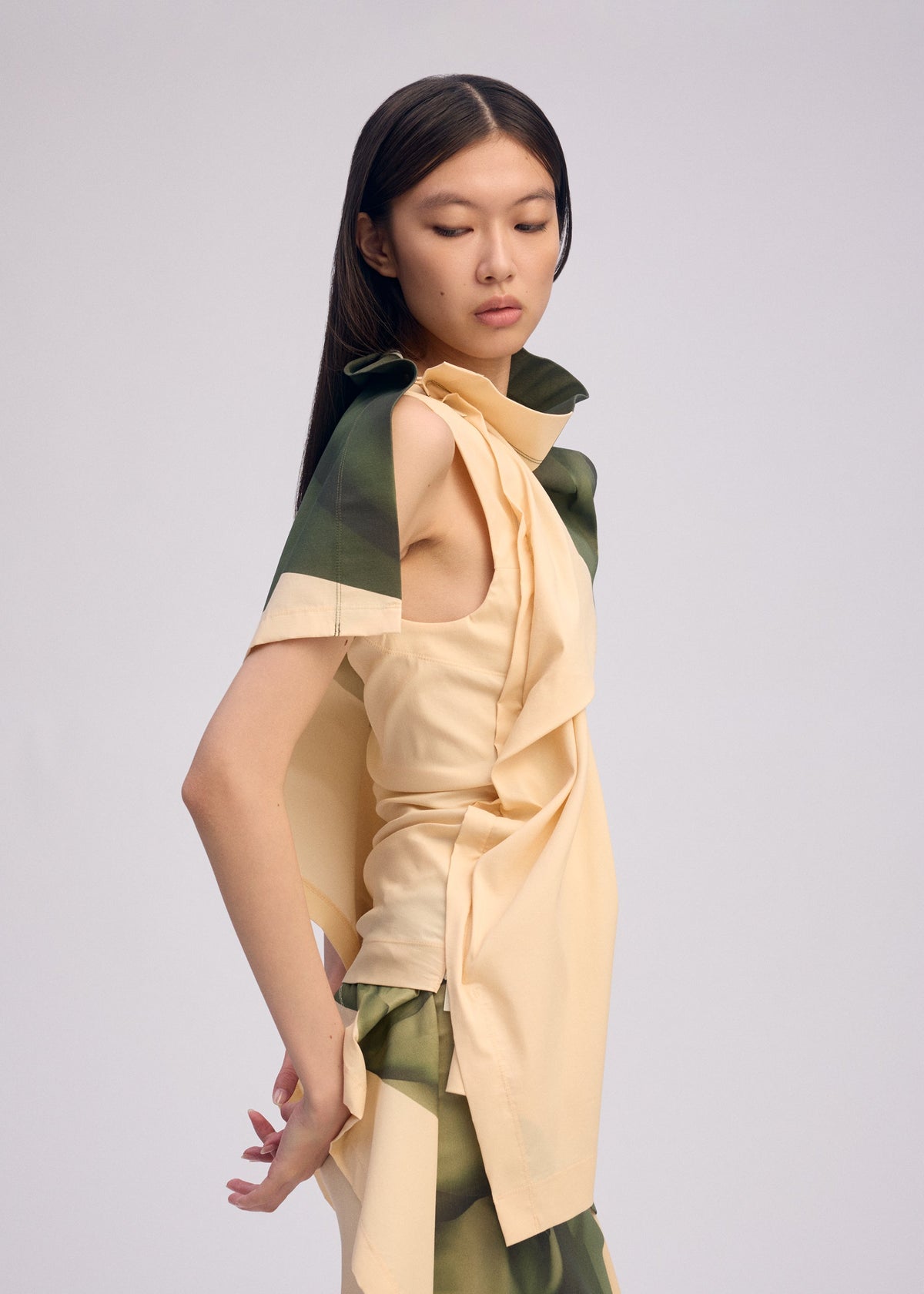 A woman with long, straight dark hair models ISSEY MIYAKE’s BODY AT PLAY: a sleeveless beige top with green accents and matching ribbed stretch skirt, posing sideways against a plain light background. view 6 of 6