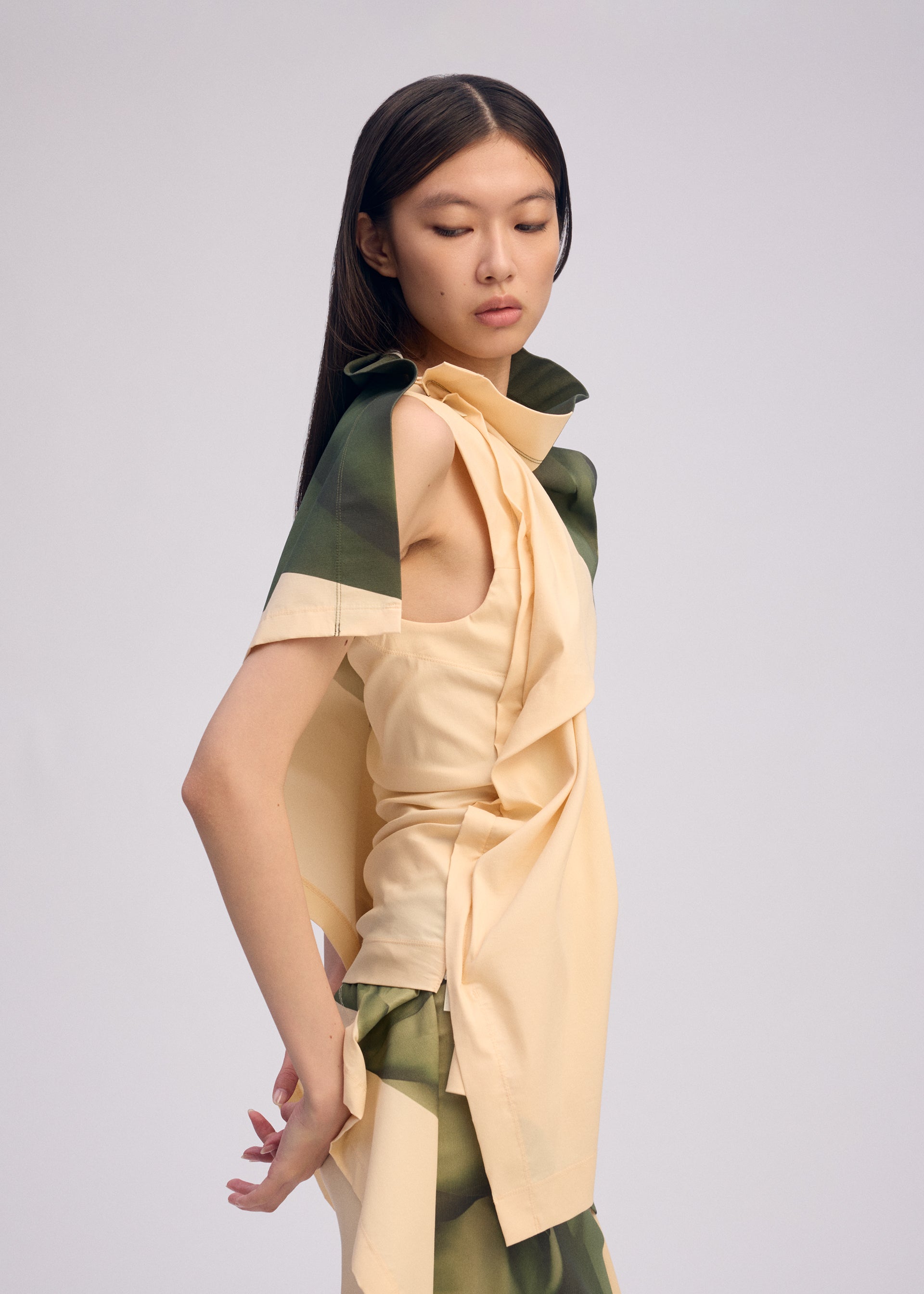 Zoomed view A woman with long, straight dark hair models ISSEY MIYAKE’s BODY AT PLAY: a sleeveless beige top with green accents and matching ribbed stretch skirt, posing sideways against a plain light background.