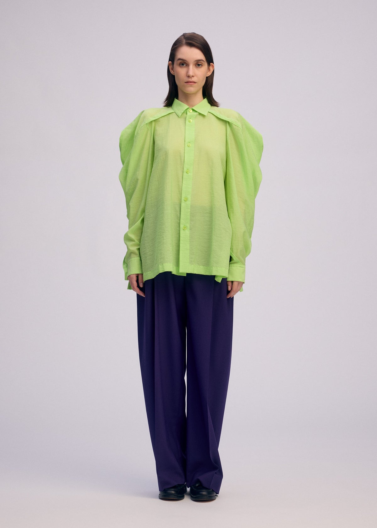 A person faces forward in a sheer, bright green ISSEY MIYAKE R SHIRT SHEER with voluminous sleeves over a dark top, paired with loose dark blue pants and black shoes, set against a plain light background. view 5 of 9