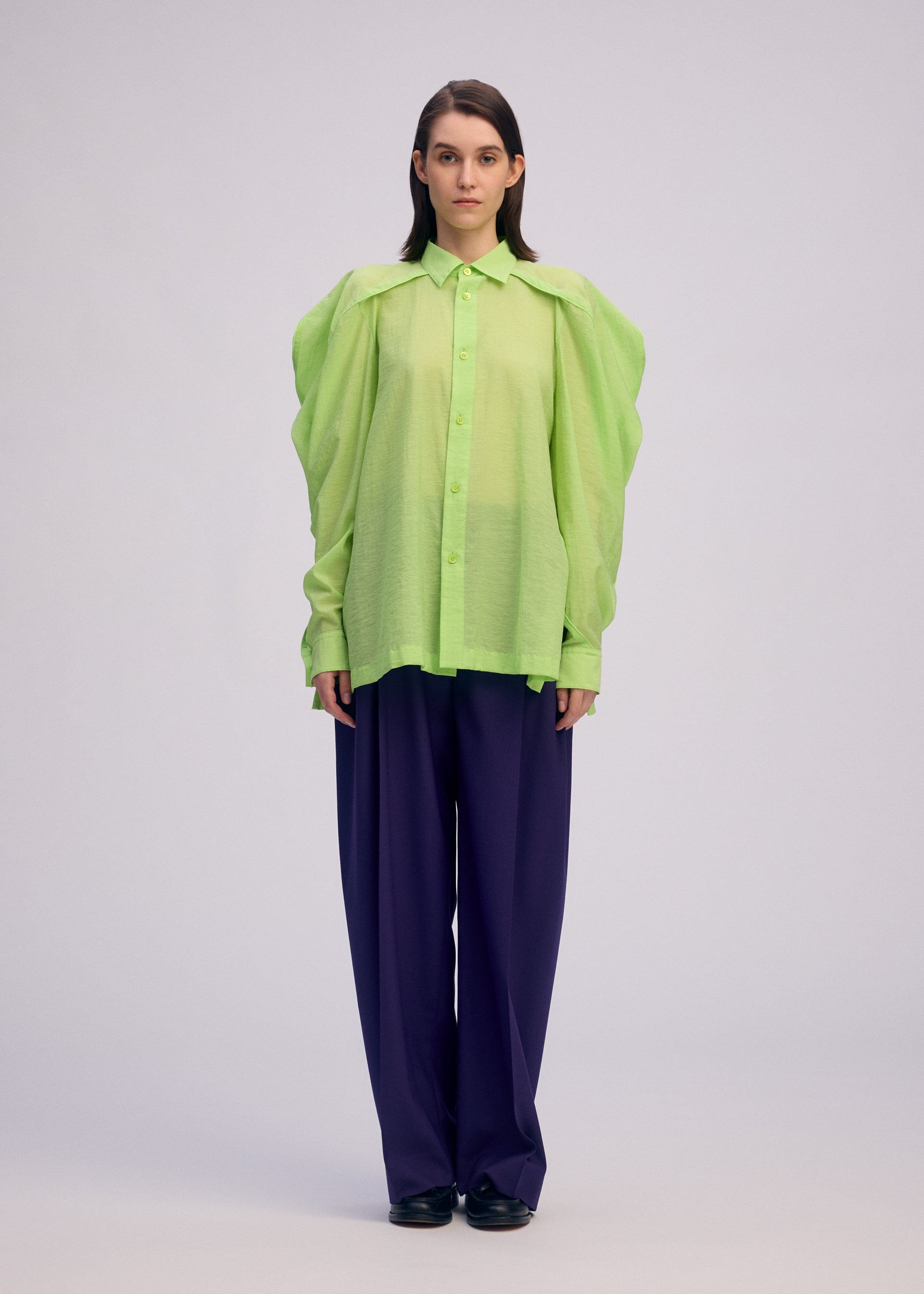 Zoomed view A person faces forward in a sheer, bright green ISSEY MIYAKE R SHIRT SHEER with voluminous sleeves over a dark top, paired with loose dark blue pants and black shoes, set against a plain light background.