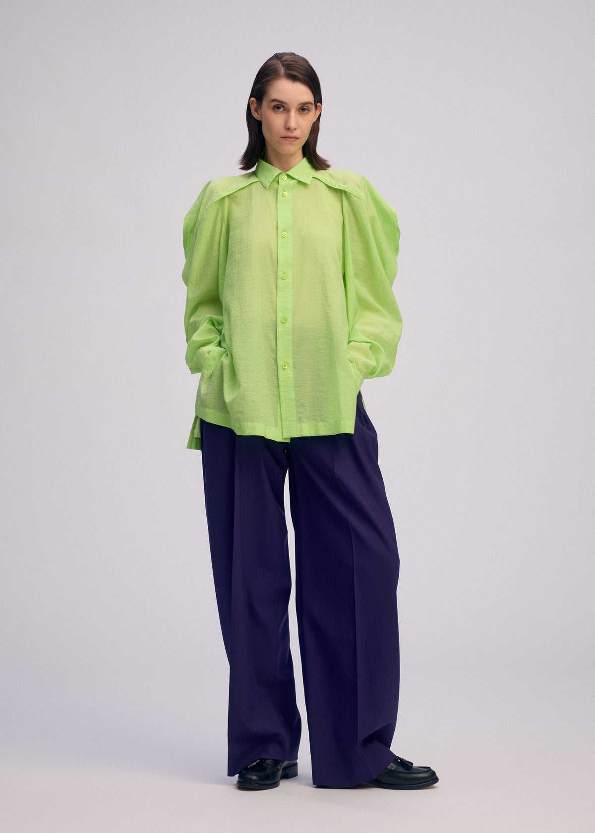 A woman with straight brown hair wears the ISSEY MIYAKE R SHIRT SHEER, a loose, bright green button-up with puffed sleeves, paired with wide-leg dark purple trousers and black shoes. She stands hands-in-pockets against a plain light background. view 6 of 9