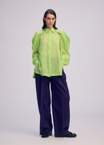 A woman with straight brown hair wears the ISSEY MIYAKE R SHIRT SHEER, a loose, bright green button-up with puffed sleeves, paired with wide-leg dark purple trousers and black shoes. She stands hands-in-pockets against a plain light background.