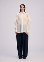 A person stands facing forward, wearing the ISSEY MIYAKE R SHIRT SHEER—an airy white button-up with oversized sleeves—paired with loose black pants against a plain, light background.