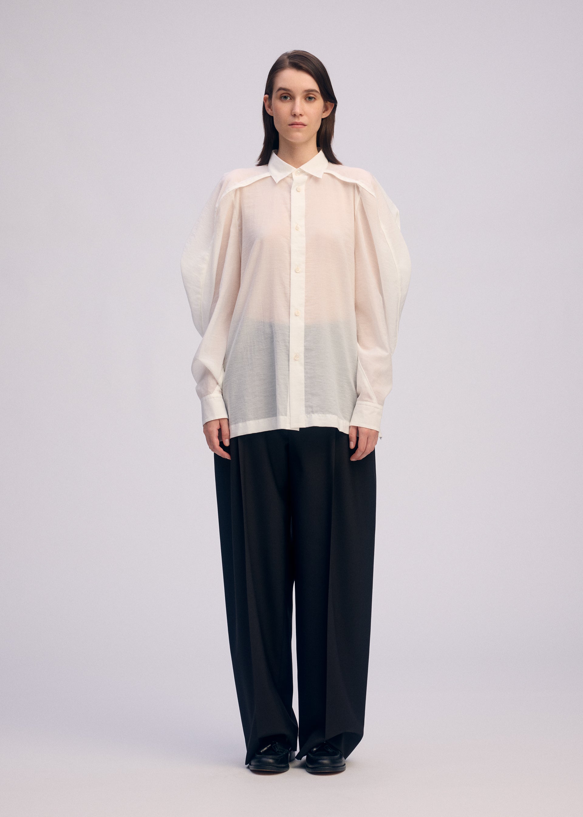 Zoomed view A person stands facing forward, wearing the ISSEY MIYAKE R SHIRT SHEER—an airy white button-up with oversized sleeves—paired with loose black pants against a plain, light background.