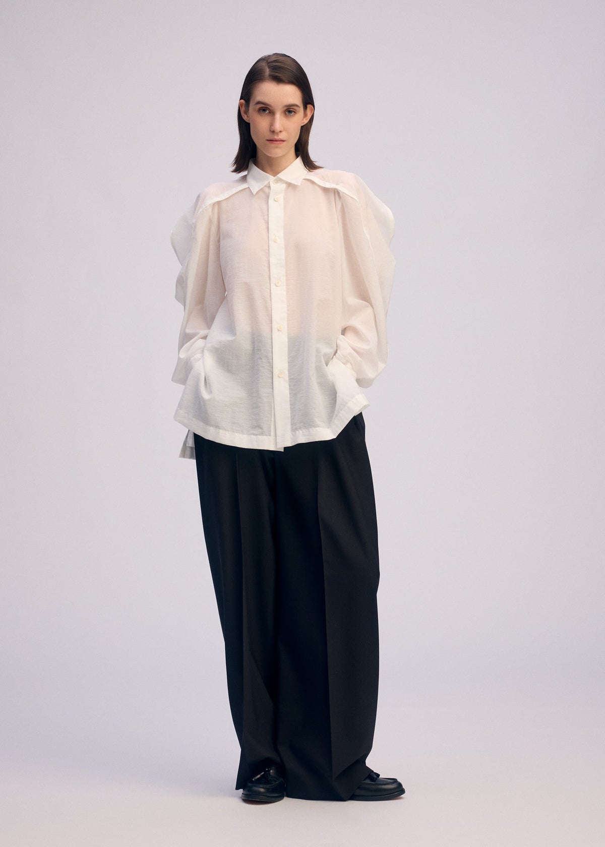 Against a plain background, a person with straight, dark hair and a neutral expression wears the ISSEY MIYAKE R SHIRT SHEER—an oversized white button-up with voluminous sleeves—paired with wide-leg black trousers and black shoes. view 8 of 9