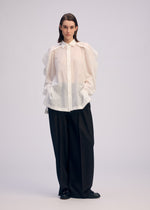 Against a plain background, a person with straight, dark hair and a neutral expression wears the ISSEY MIYAKE R SHIRT SHEER—an oversized white button-up with voluminous sleeves—paired with wide-leg black trousers and black shoes.
