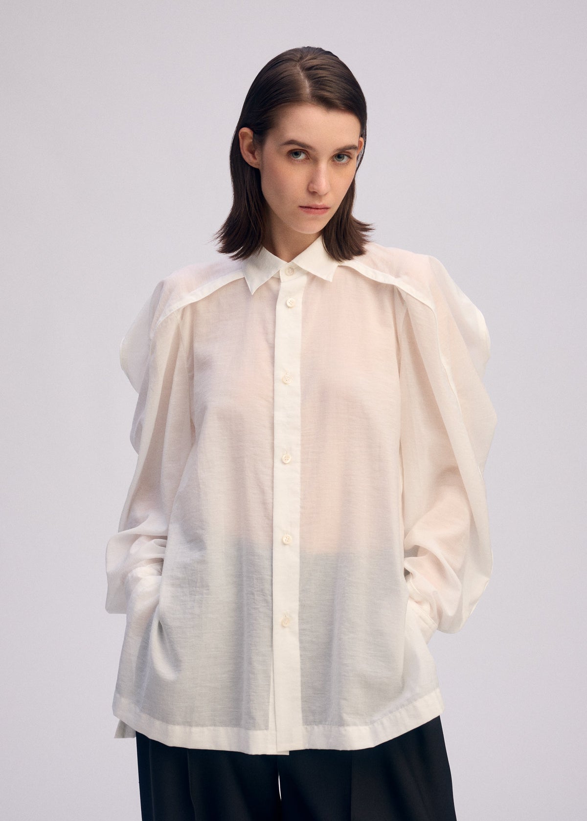 A person with straight, shoulder-length brown hair wears the ISSEY MIYAKE R SHIRT SHEER, a white, button-up blouse with voluminous sleeves and a tissue-like texture, paired with black pants against a plain light background. view 9 of 9