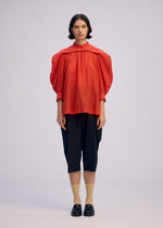 A person faces forward in a bright red ISSEY MIYAKE R SHIRT SHEER with voluminous, tissue-like sleeves, black knee-length culottes, beige socks, and black loafers against a plain light background.