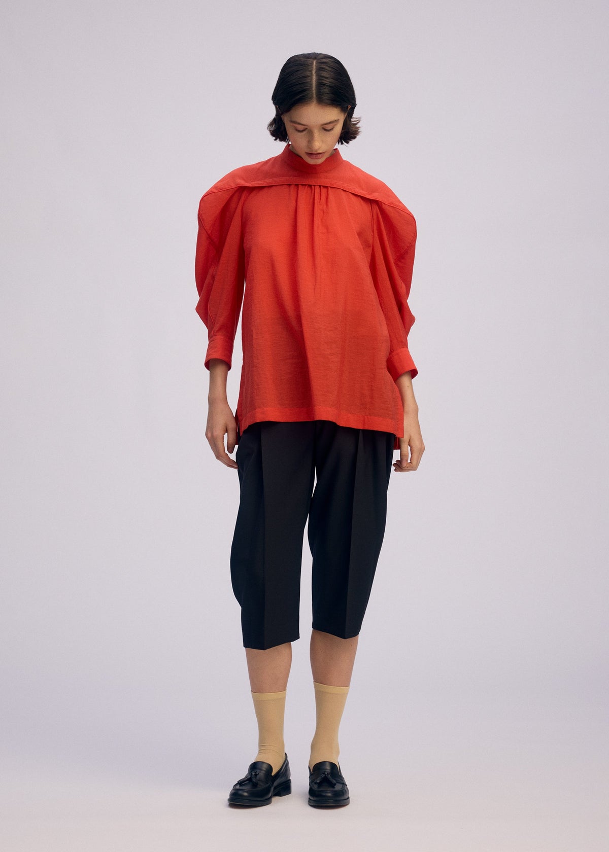 A person stands against a light background, wearing the ISSEY MIYAKE R SHIRT SHEER—a bright red, tissue-textured standing collar shirt—paired with black knee-length shorts, beige socks, black loafers, and sporting short dark hair. view 6 of 8