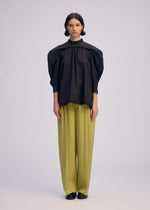 A person stands facing forward, dressed in the ISSEY MIYAKE R SHIRT SHEER—a loose black blouse with puffed sleeves and textured fabric—paired with wide-leg light green trousers and black shoes, against a plain light background.