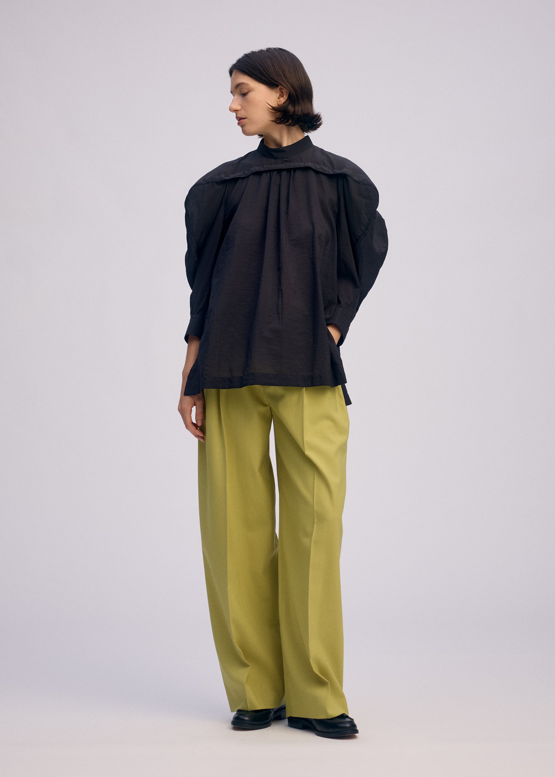Zoomed view A person stands against a plain background wearing the ISSEY MIYAKE R SHIRT SHEER, a black standing collar shirt with wide sleeves, paired with wide-leg light green trousers and black shoes. They look to the side with one hand in a pocket.