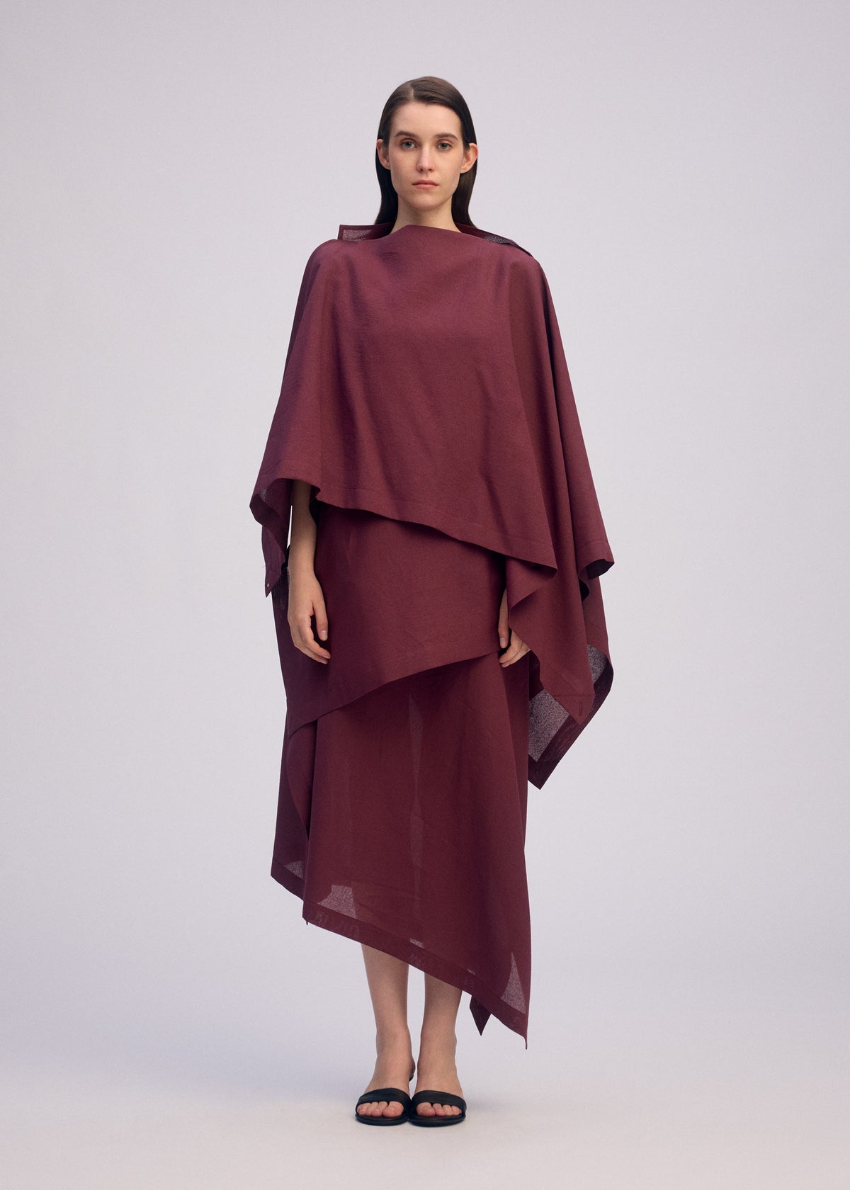 A woman faces forward in a loose, layered maroon ALL SQUARES dress by ISSEY MIYAKE with asymmetrical hems. She has straight brown hair, minimal makeup, and wears black sandals, standing barefoot against a plain light gray background. view 5 of 8