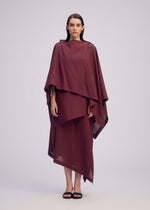 A woman faces forward in a loose, layered maroon ALL SQUARES dress by ISSEY MIYAKE with asymmetrical hems. She has straight brown hair, minimal makeup, and wears black sandals, standing barefoot against a plain light gray background.