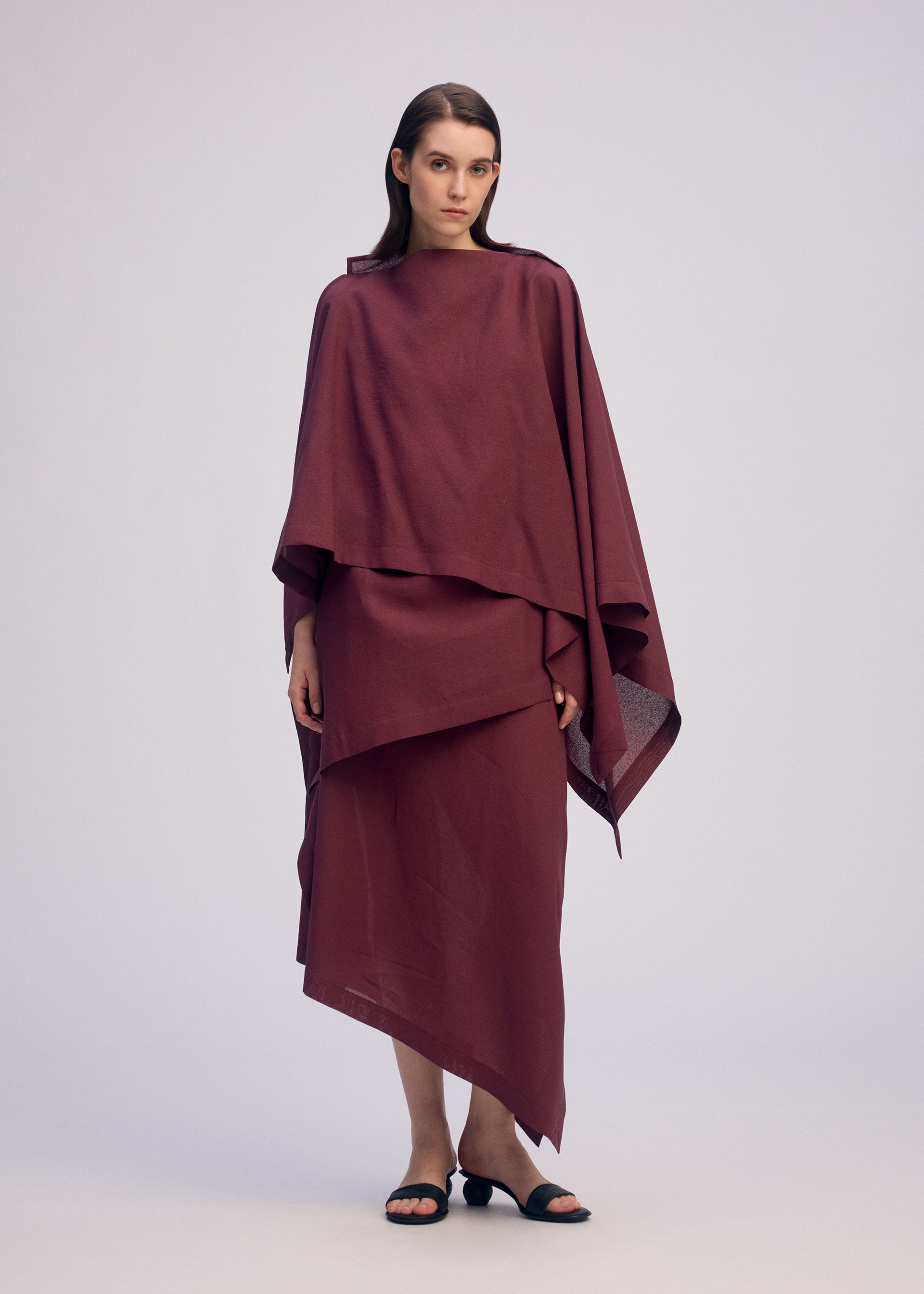 Zoomed view A woman with straight, center-parted hair and a neutral expression wears ISSEY MIYAKE's ALL SQUARES, a loose, layered burgundy outfit featuring asymmetrical hemlines and a draped top, standing against a plain background.