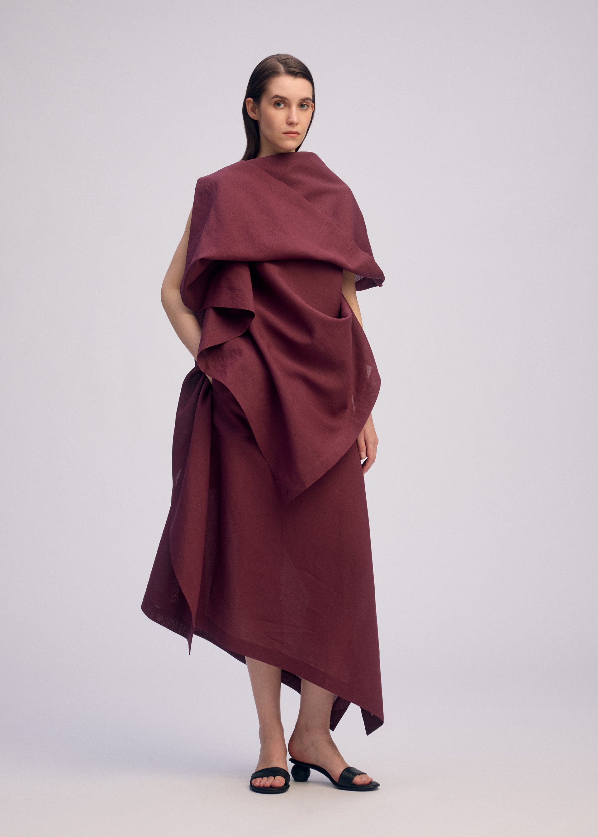 Zoomed view A woman with straight brown hair stands against a plain background in the ISSEY MIYAKE ALL SQUARES dress, featuring a burgundy draped top with layered, asymmetrical fabric. She completes her look with black heeled sandals.