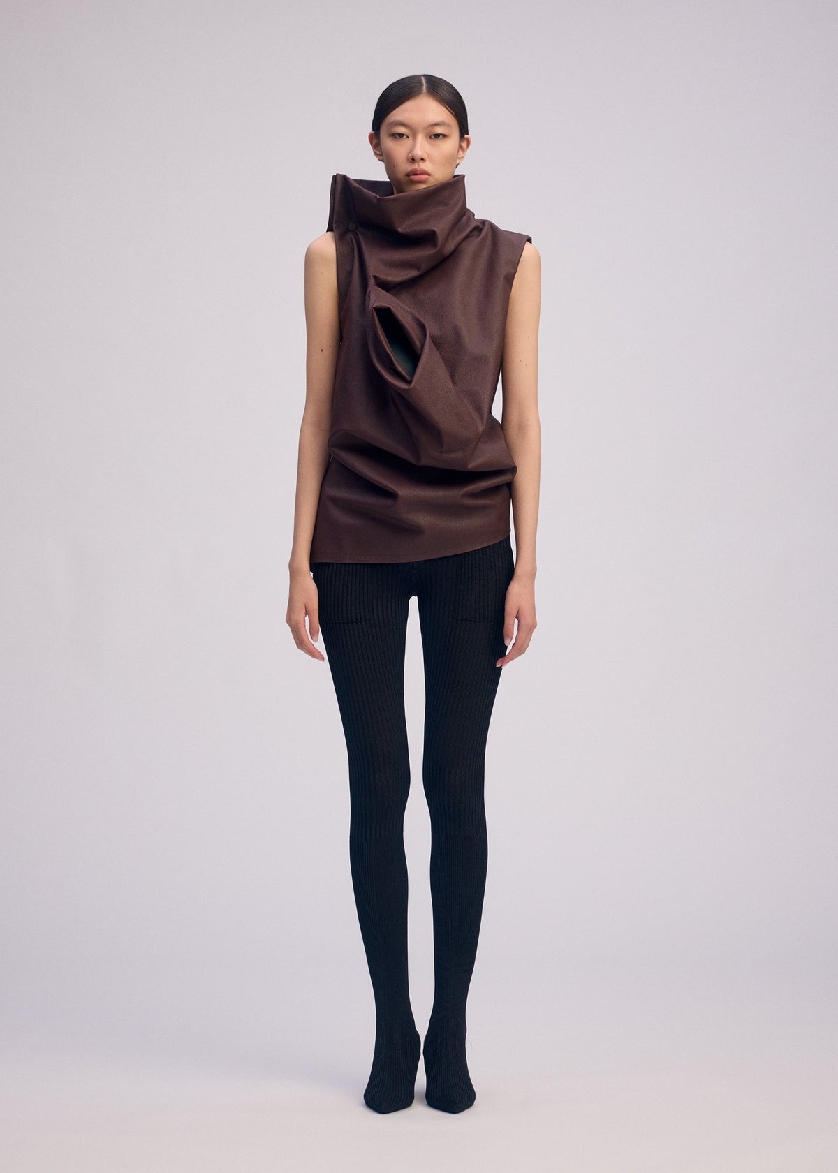 A person stands against a plain backdrop wearing a sleeveless, sculptural artificial leather top with large folds and a circular cutout, paired with black ribbed leggings and sleek ISSEY MIYAKE PEU FORM³ shoes. view 4 of 7