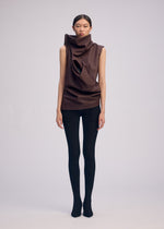 A person stands against a plain backdrop wearing a sleeveless, sculptural artificial leather top with large folds and a circular cutout, paired with black ribbed leggings and sleek ISSEY MIYAKE PEU FORM³ shoes.