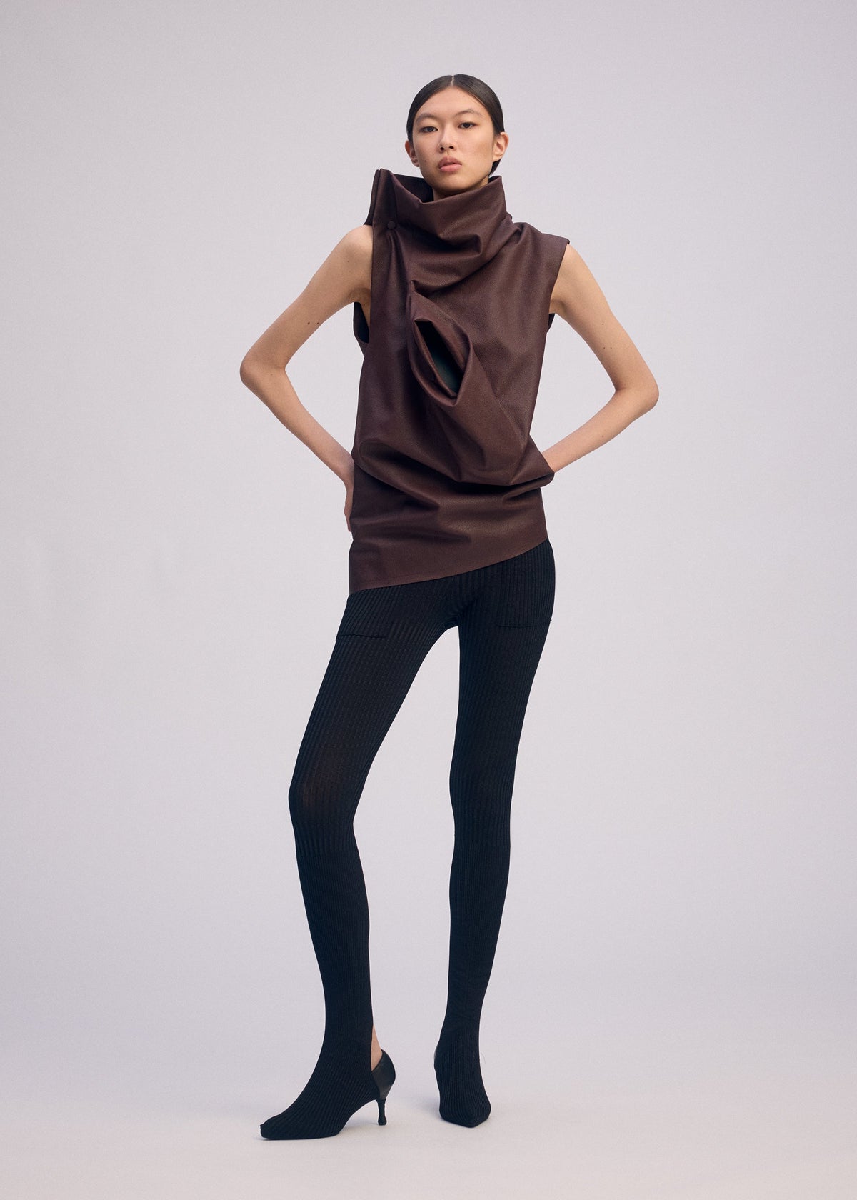 A woman stands against a plain background wearing an artificial leather top with a sculptural draped collar and cutout, paired with black textured leggings and ISSEY MIYAKE PEU FORM³ shoes. view 5 of 7