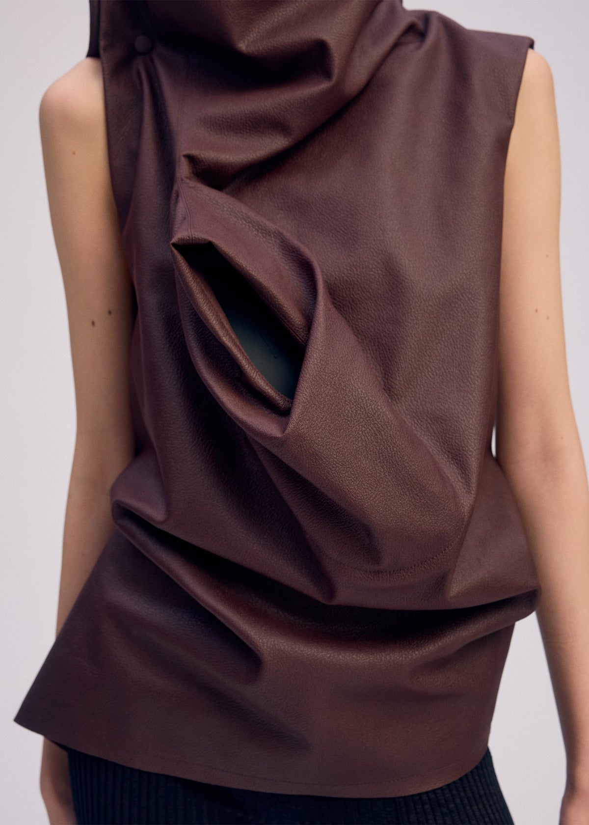 A person wears ISSEY MIYAKE's PEU FORM³—a sleeveless, dark brown faux leather top with a sculptural front fold and opening—paired with black ribbed pants. Part of a Camper collaboration. Image is cropped at shoulders and waist. view 7 of 7