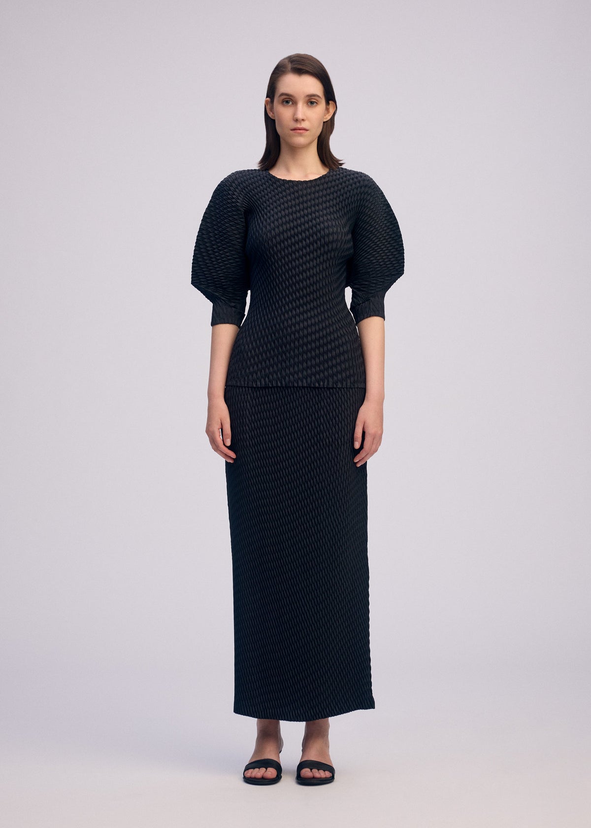 A woman with straight brown hair stands facing forward in ISSEY MIYAKE’s SLEEK PLEATS—an elegant pleated satin top with elbow-length sleeves and a matching long skirt, paired with black sandals, set against a plain light background. view 5 of 7