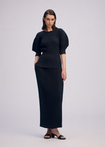 A woman with shoulder-length hair poses relaxed against a plain background, wearing ISSEY MIYAKE’s SLEEK PLEATS set: a round-neck, short-sleeved top with structured shoulders and a matching long skirt, paired with black sandals.