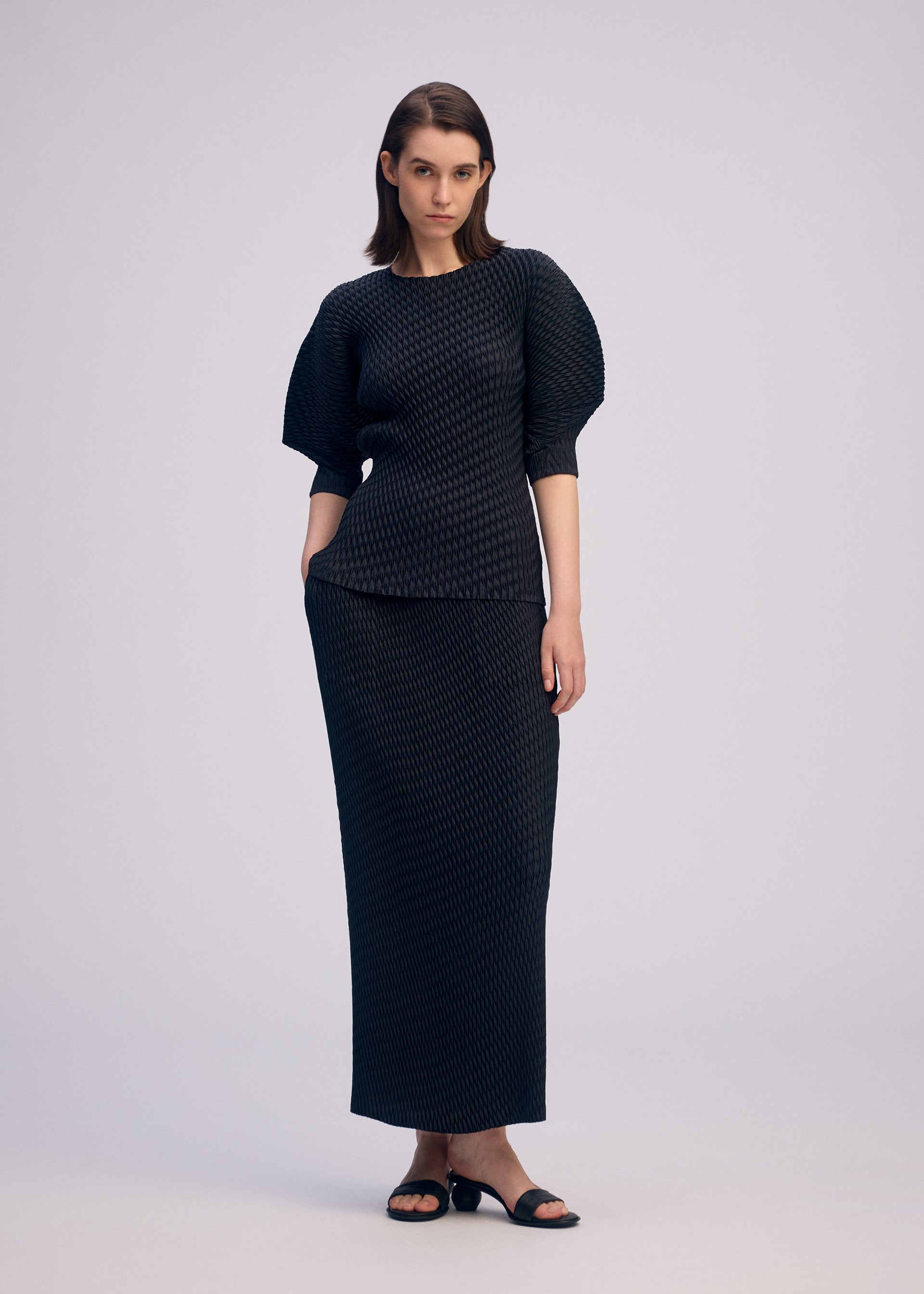 Zoomed view A woman with shoulder-length hair poses relaxed against a plain background, wearing ISSEY MIYAKE’s SLEEK PLEATS set: a round-neck, short-sleeved top with structured shoulders and a matching long skirt, paired with black sandals.
