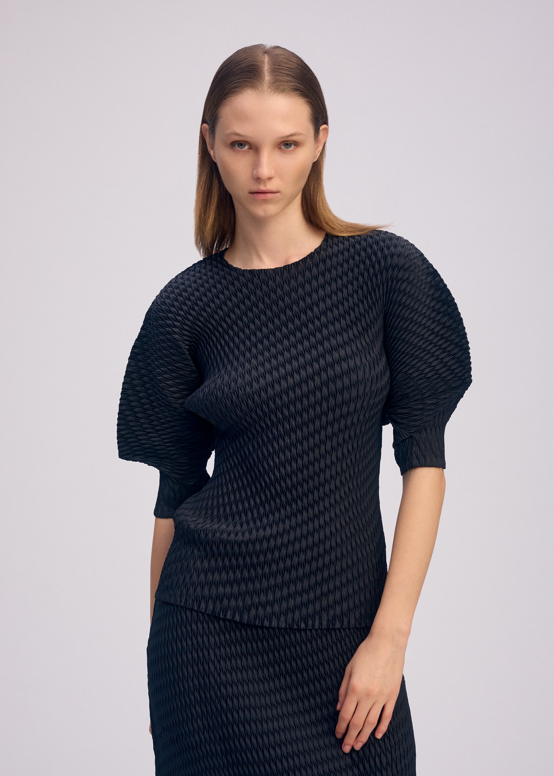 Zoomed view A woman with straight, light brown hair models the ISSEY MIYAKE SLEEK PLEATS set—glossy blouse with voluminous sleeves and matching skirt—posing against a plain, light background.