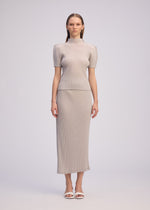 A woman stands facing forward, wearing the ISSEY MIYAKE BOX SHOULDER PLEATS outfit—a light beige ribbed knit set with a high-neck top and box shoulder silhouette, paired with a matching calf-length skirt and white open-toe sandals.