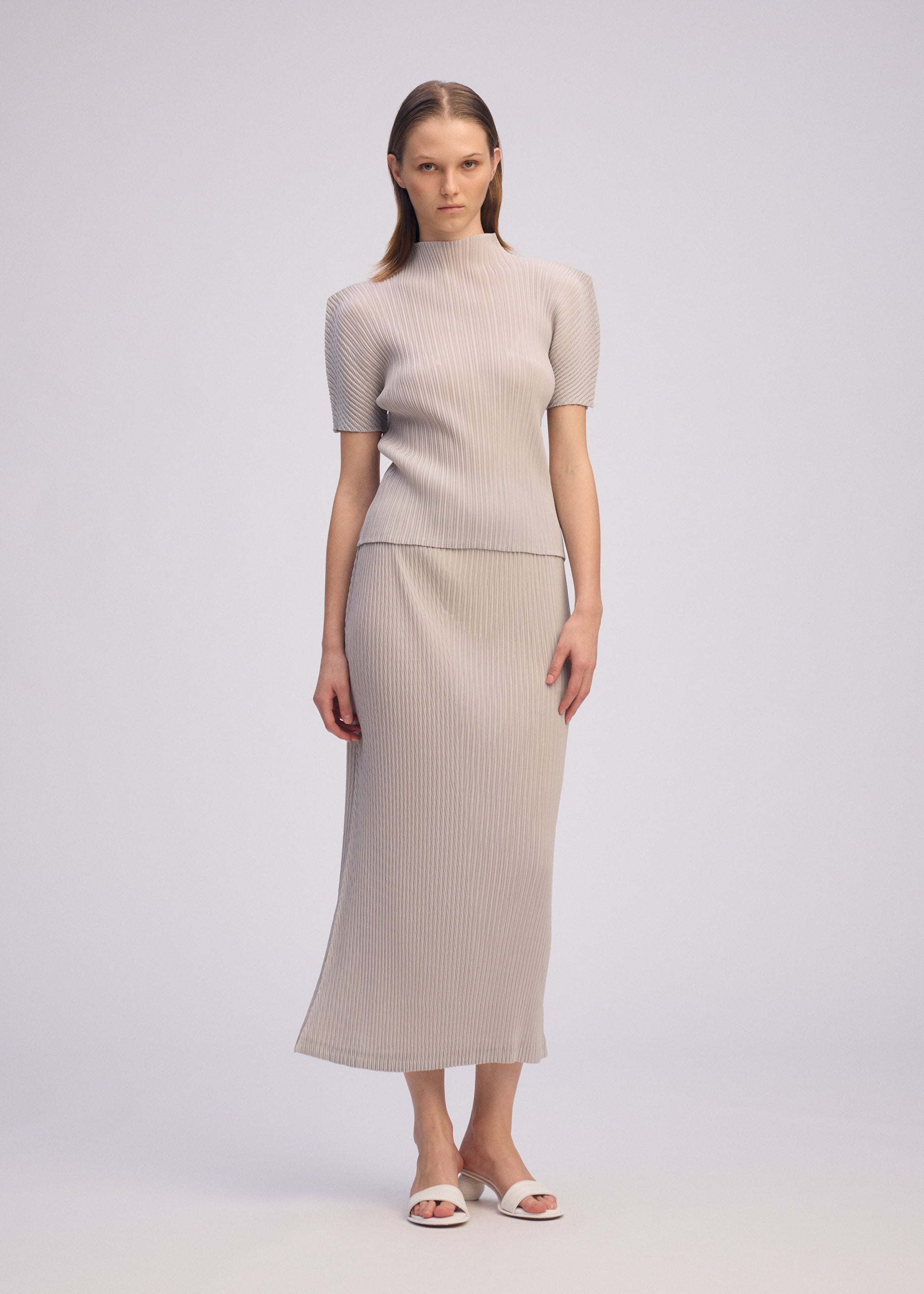 Zoomed view A woman with straight hair and a neutral expression wears the ISSEY MIYAKE BOX SHOULDER PLEATS set—a light gray, ribbed high-neck top and matching long skirt—paired with white open-toe sandals against a plain background.