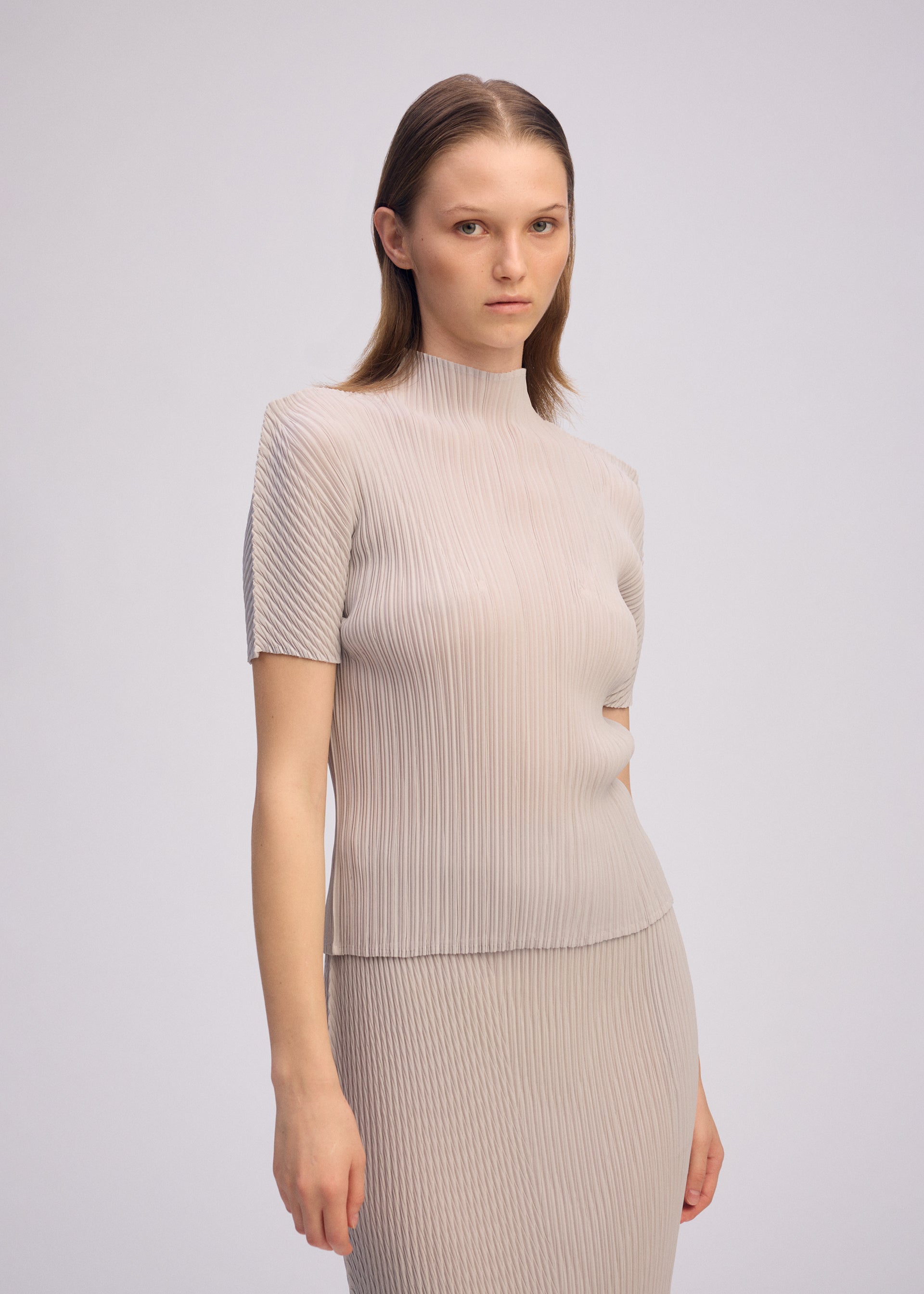 Zoomed view A woman with straight brown hair wears the ISSEY MIYAKE BOX SHOULDER PLEATS set—a light beige, high-neck top and matching pleated skirt—standing against a plain background, gazing neutrally at the camera.
