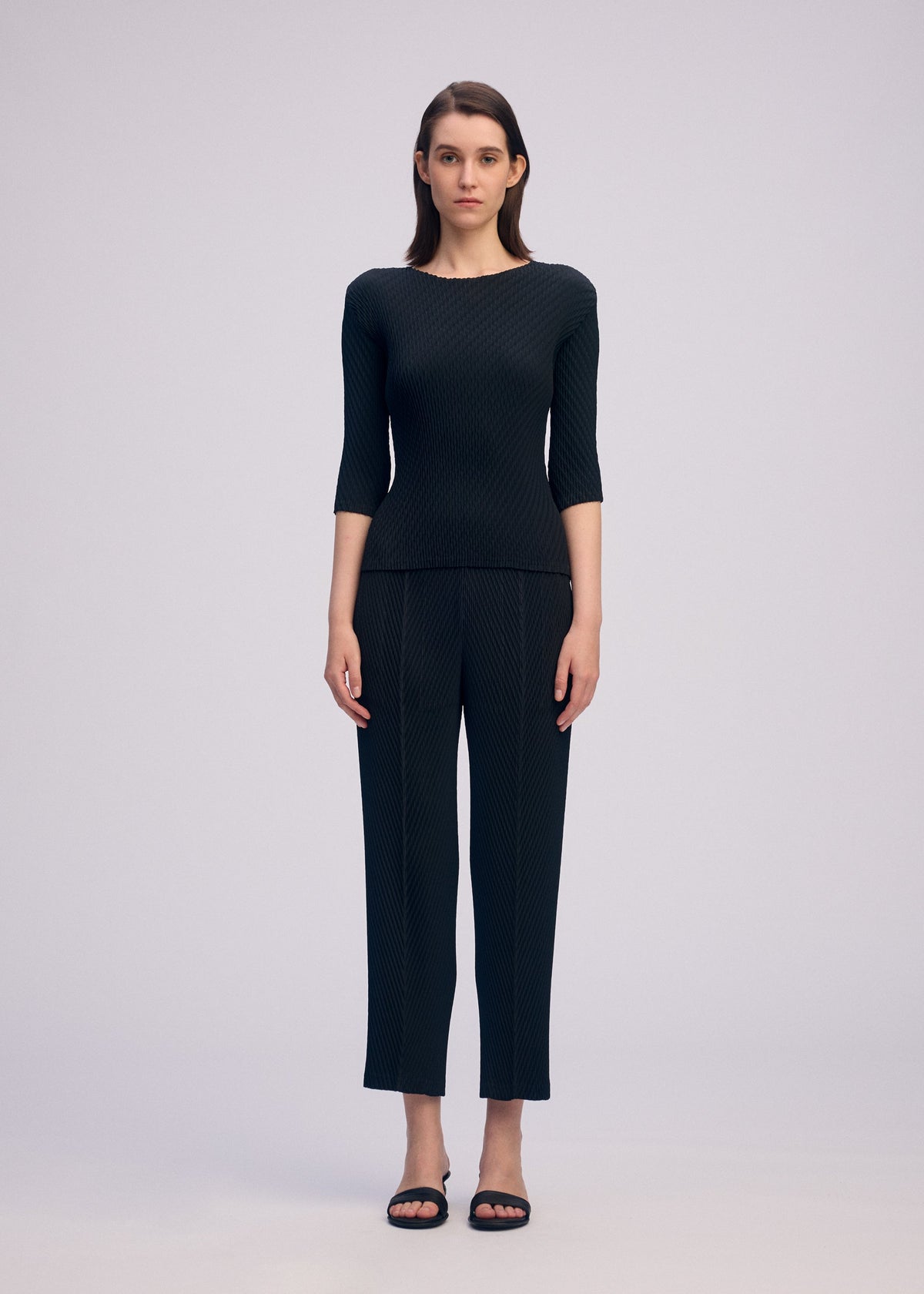 A woman stands facing forward against a plain background, wearing the ISSEY MIYAKE RHOMBUS PLEATS black round-neck top and matching straight-leg pants. She has straight brown hair, black open-toed sandals, and presents a sleek silhouette. view 5 of 6