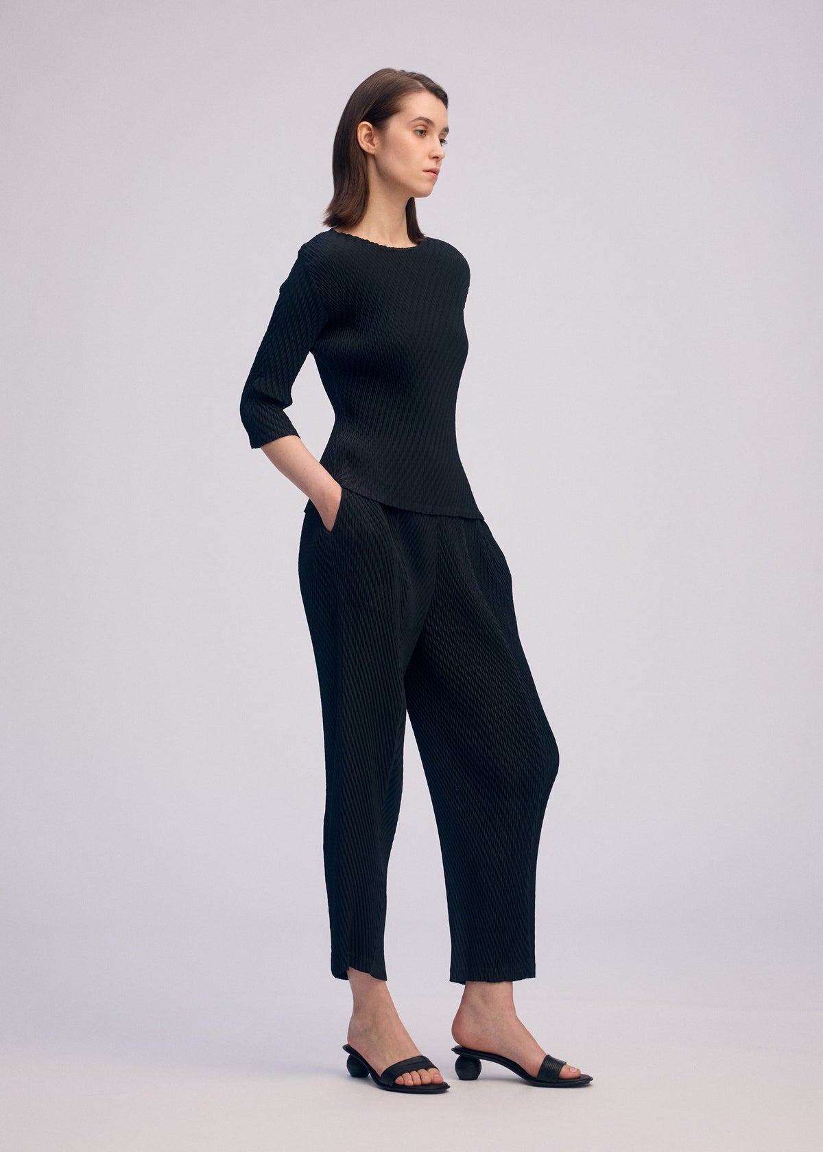 A woman poses in ISSEY MIYAKE’s RHOMBUS PLEATS set—a black round-neck top with three-quarter sleeves and matching wide-leg pants—paired with black open-toe heels against a plain light background. view 6 of 6