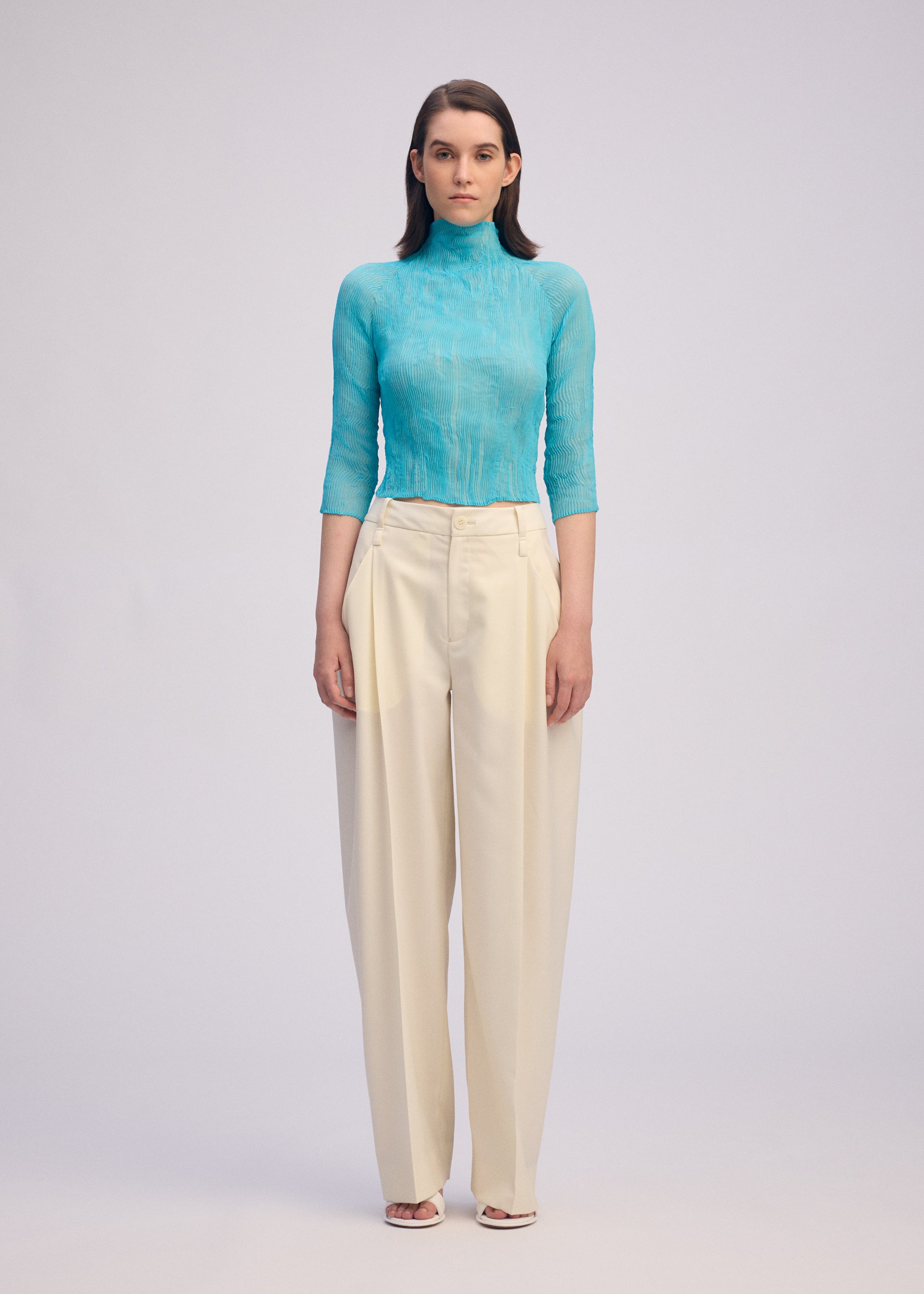 Zoomed view A woman stands facing forward, wearing the ISSEY MIYAKE CHIFFON TWIST 2 sheer turquoise high-neck top with three-quarter sleeves and high-waisted wide-leg cream trousers. She has straight shoulder-length hair and open-toed shoes.