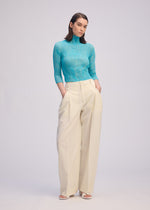 A person with straight, shoulder-length hair stands against a plain background, wearing ISSEY MIYAKE's CHIFFON TWIST 2—a sheer high-neck turquoise top—paired with high-waisted, wide-leg cream pleated trousers and white open-toe shoes.