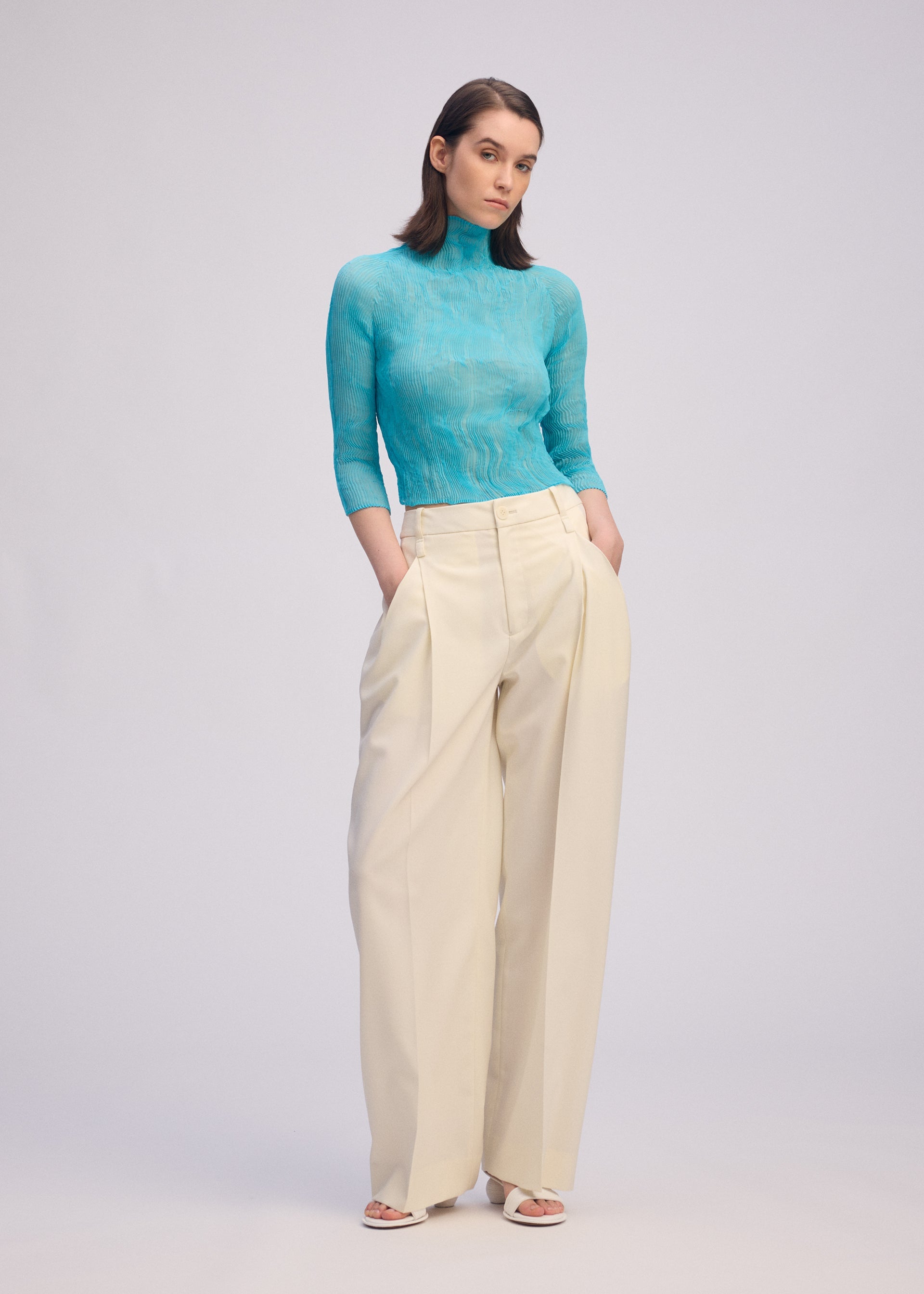 Zoomed view A person with straight, shoulder-length hair stands against a plain background, wearing ISSEY MIYAKE's CHIFFON TWIST 2—a sheer high-neck turquoise top—paired with high-waisted, wide-leg cream pleated trousers and white open-toe shoes.