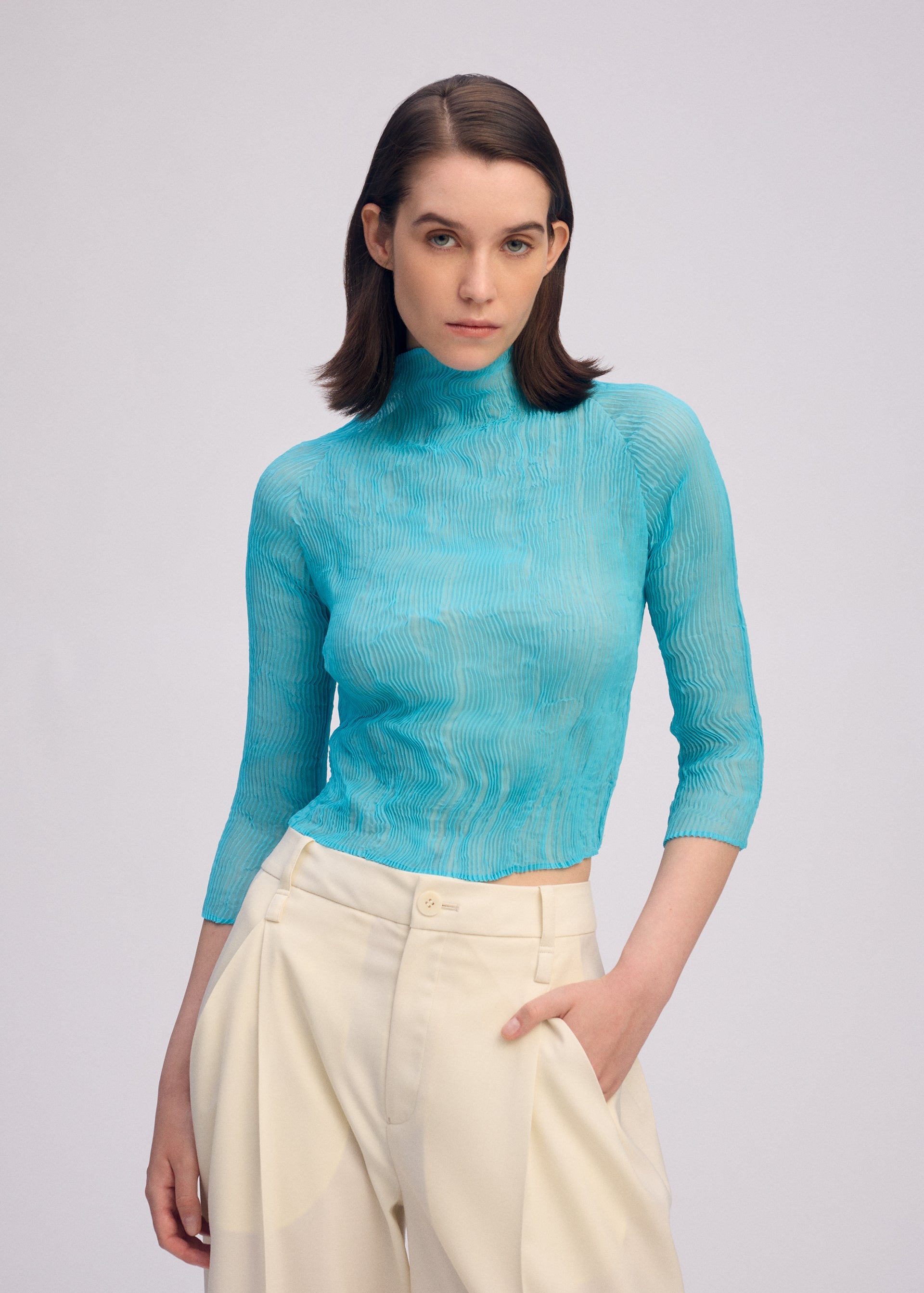 Zoomed view A woman with straight brown hair wears the ISSEY MIYAKE CHIFFON TWIST 2, a sheer high-neck light blue top with high-waisted cream pleated recycled polyester trousers, posing with one hand in her pocket against a plain background.