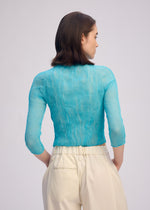 A woman with straight brown hair, seen from behind, wears the ISSEY MIYAKE CHIFFON TWIST 2—a sheer turquoise high-neck top with three-quarter sleeves and pleated high-waisted cream trousers—standing against a plain light background.