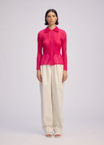 A woman with short, dark hair faces forward against a plain background, wearing the ISSEY MIYAKE CHIFFON TWIST 1 bright pink pleated zip-up cardigan with a collar, paired with loose high-waisted cream trousers and white open-toe shoes.